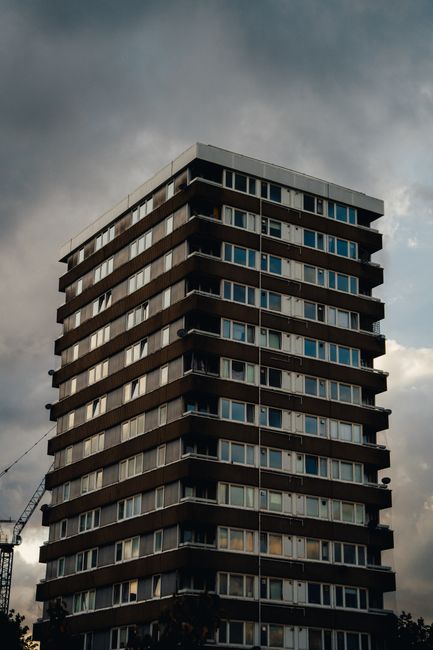 A tall, multi-storey residential building with several floors featuring horizontal bands of dark material and large rectangular windows, some of which are open. The building is set against a cloudy sk