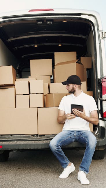 A man wearing a white T-shirt, black cap, and jeans is sitting on the edge of an open van with its rear doors wide open. Inside the van, there are various stacked cardboard boxes of different sizes, s