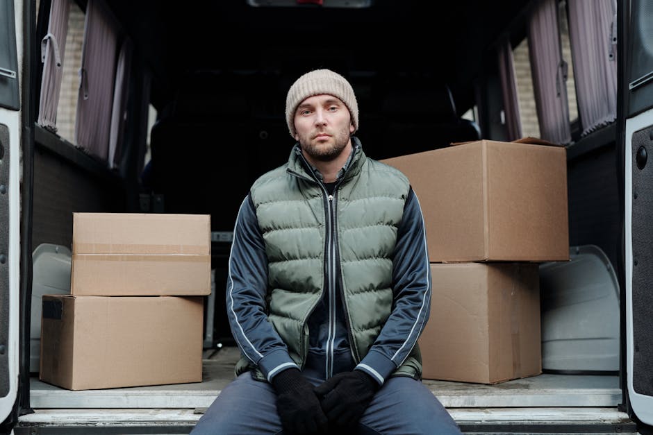A man wearing a white T-shirt, black cap, and jeans is sitting on the edge of an open van with its rear doors wide open. Inside the van, there are various stacked cardboard boxes of different sizes, s
