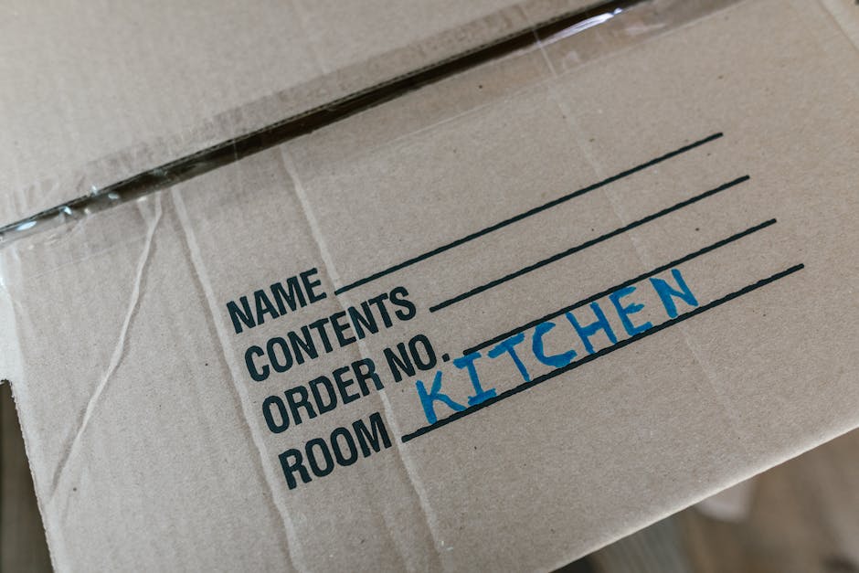 A cardboard box labeled 'SUMMER CLOTHS' with additional labels indicating 'MEDIUM' size and a white tape roll on top, situated on a wooden floor. Behind it, another box with similar 'SUMMER CLOTHS' la